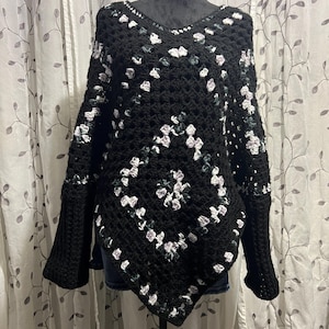 May include: A black crochet poncho with a V-neck and bell sleeves. The poncho features a floral pattern in white and light purple. The garment is displayed on a mannequin.