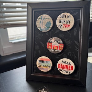 May include: A black rectangular frame containing five circular pin-back buttons. The buttons feature various designs including text that reads: "FUND LIBRARIES", "BURY ME WITH MY TBR", "FUND THE DATA CENTER", "FREND of the GLENSIDE Library", and "READ BANNED BOOKS".