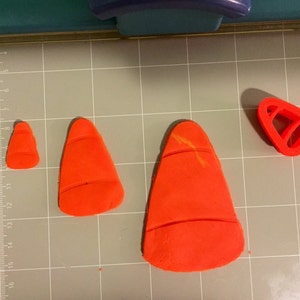 May include: Three orange fondant cones of different sizes, and a red plastic cone-shaped cookie cutter on a gray grid surface.