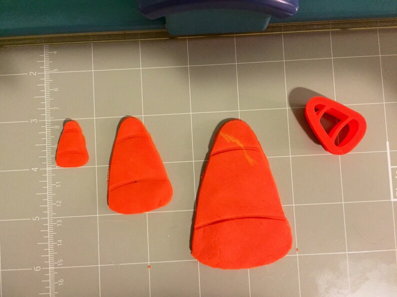 May include: Three orange fondant cones of different sizes, and a red plastic cone-shaped cookie cutter on a gray grid surface.