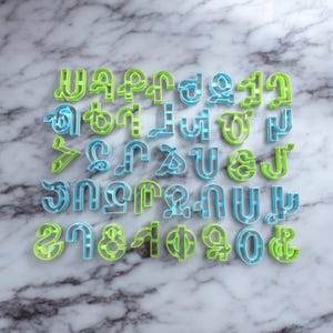 Puede incluir: Un juego de 26 cortadores de galletas con forma de letras del alfabeto en color azul claro y verde. Los cortadores están dispuestos en orden alfabético sobre una superficie de mármol.