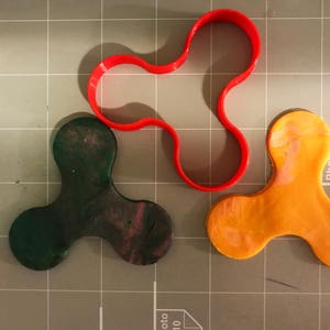 May include: A red plastic cookie cutter shaped like a three-pronged fidget spinner, with two finished clay fidget spinners, one green and purple and the other orange.