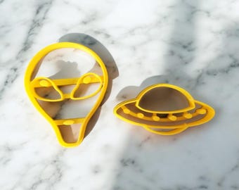 May include: Two yellow plastic cookie cutters, one shaped like an alien head and the other shaped like a flying saucer.