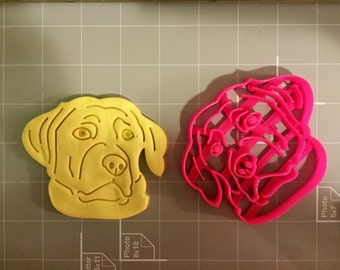 Könnte beinhalten: Zwei Hundeförmige Ausstecher, einer gelb und einer rosa. Der gelbe Ausstecher ist eine einfache Umrandung eines Hundekopfes, während der rosa Ausstecher ein detaillierteres Design mit Linien und Streifen hat.
