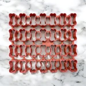May include: A red plastic cookie cutter with 24 bone-shaped cutouts. The cookie cutter is designed to create multiple cookies at once.