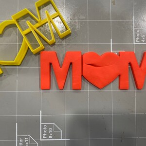 May include: A red fondant "MOM" with a heart in the middle, next to a yellow plastic cookie cutter of the same word.
