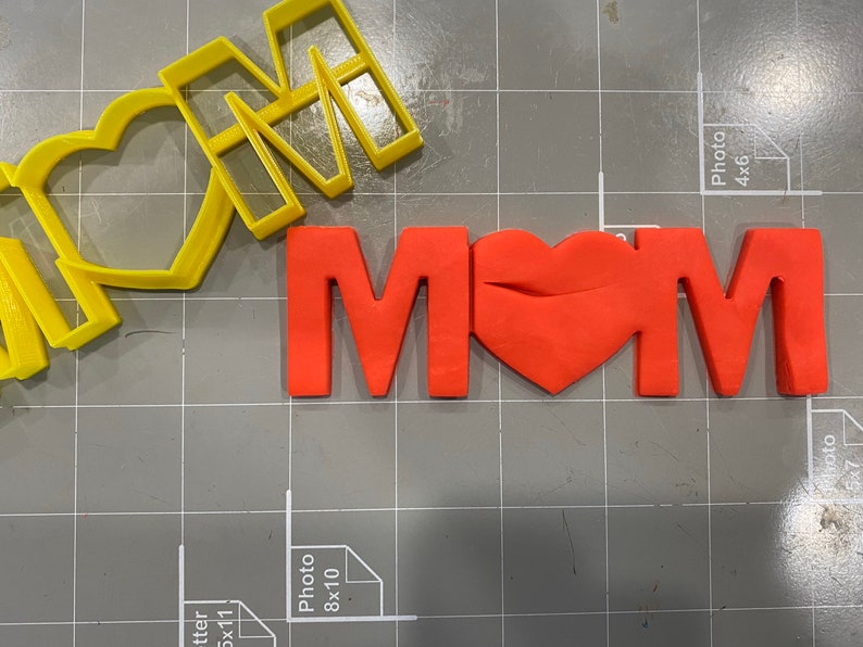 May include: A red fondant "MOM" with a heart in the middle, next to a yellow plastic cookie cutter of the same word.