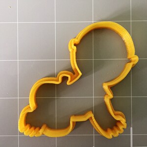 May include: A yellow plastic cookie cutter shaped like a baby. The cutter has a rounded head and a body with arms and legs.