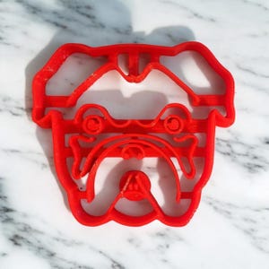 Puede incluir: Cortador de galletas de bulldog de plástico rojo. El cortador tiene forma de cabeza de bulldog con una cara detallada.