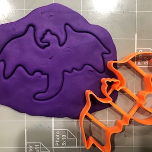 May include: A purple fondant with a dragon cookie cutter pressed into it. A separate orange plastic dragon cookie cutter is shown next to the fondant.