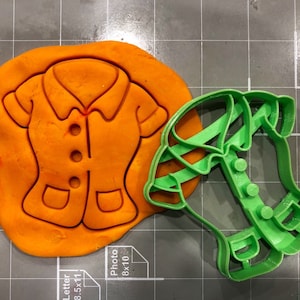 Pode incluir: Um cortador de biscoitos de plástico verde em forma de camisa com colarinho, botões e bolsos. O cortador de biscoitos está em cima de um pedaço de argila de modelar laranja.