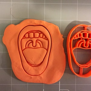 May include: A close-up of an orange plastic cookie cutter shaped like an open mouth with teeth and a tongue. The cookie cutter is on a grey surface.