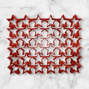 Puede incluir: Un juego de 24 cortadores de galletas en forma de estrella roja. Los cortadores están dispuestos en un patrón de cuadrícula, con cinco filas de cuatro cortadores cada uno.