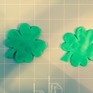 May include: Two green four-leaf clover shapes made of fondant or modeling clay. The clovers are on a gray gridded surface.