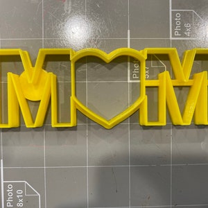 May include: Yellow plastic cookie cutter in the shape of the word "MOM" with a heart in the middle.