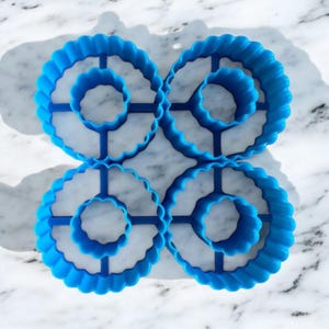 Puede incluir: Cuatro cortadores de galletas de plástico azul en forma de rosquilla con bordes festoneados. Los cortadores están dispuestos en un patrón cuadrado.