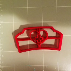 May include: Red plastic cookie cutter shaped like a nurse's hat with a heart and cross in the center.