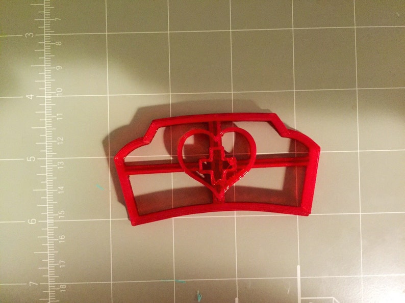 May include: Red plastic cookie cutter shaped like a nurse's hat with a heart and cross in the center.