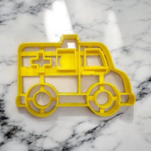 Pode incluir: Cortador de biscoitos de plástico amarelo em forma de ambulância. O cortador tem uma cruz no lado e uma janela na frente.