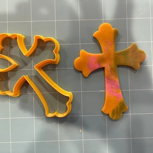 May include: Two yellow plastic cookie cutters in the shape of a cross. The cross on the left has a more intricate design with a scalloped edge. The cross on the right is a simpler design with a smooth edge.