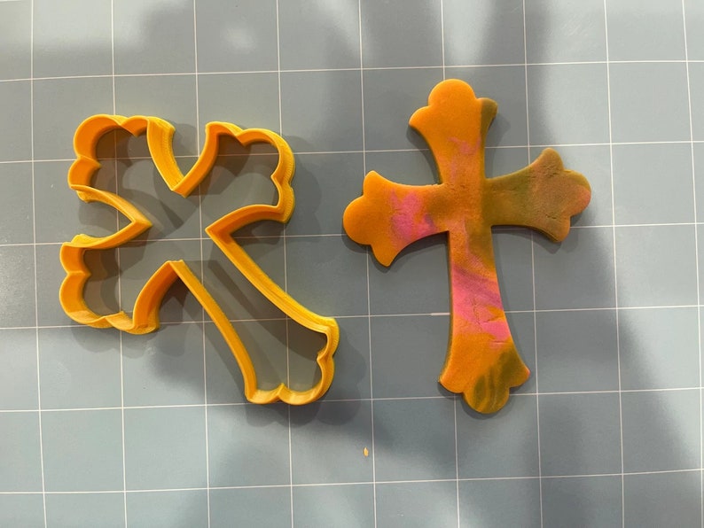 May include: Two yellow plastic cookie cutters in the shape of a cross. The cross on the left has a more intricate design with a scalloped edge. The cross on the right is a simpler design with a smooth edge.