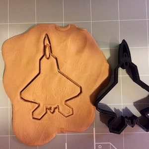 May include: A black metal cookie cutter in the shape of an F-22 Raptor fighter jet. The cutter is shown next to a piece of brown modeling clay with the jet shape cut out.