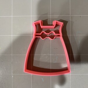 May include: Pink plastic cookie cutter shaped like a dress with a bow at the waist.