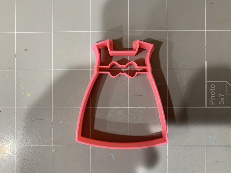 May include: Pink plastic cookie cutter shaped like a dress with a bow at the waist.