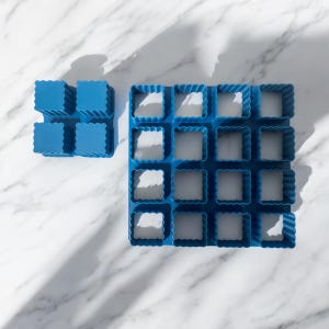 Puede incluir: Dos moldes de silicona azules para alimentos, uno con 6 cavidades cuadradas y el otro con 16. Los moldes tienen un diseño de borde ondulado. Los moldes están sobre una superficie de mármol blanco.