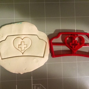 Pode incluir: Um cortador de biscoitos de plástico vermelho em forma de chapéu de enfermeira com um coração e uma cruz no centro. O cortador está ao lado de uma peça de massa de modelar branca com o mesmo desenho impresso nela.