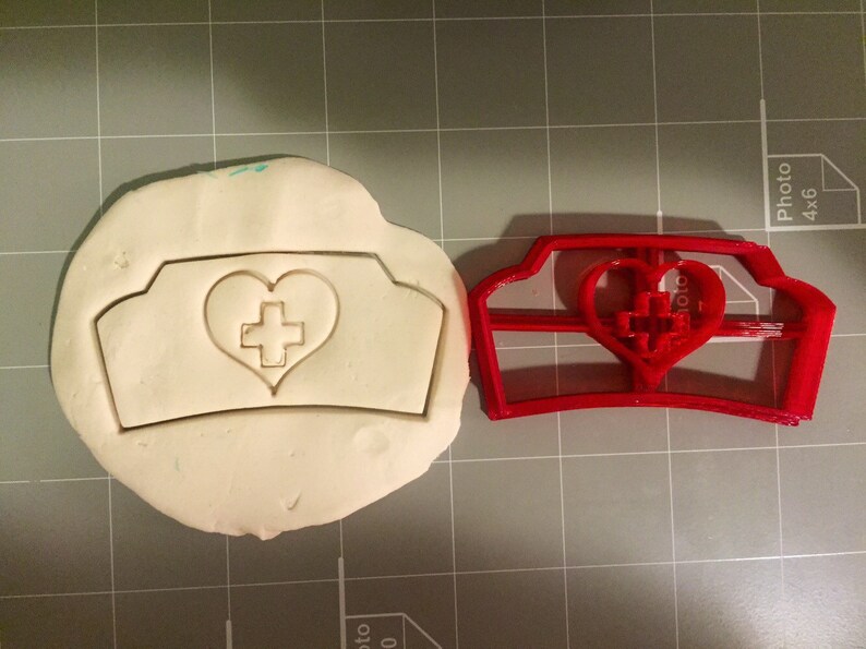 May include: A red plastic cookie cutter shaped like a nurse's hat with a heart and cross in the center. The cutter is next to a white piece of modeling clay with the same design imprinted in it.