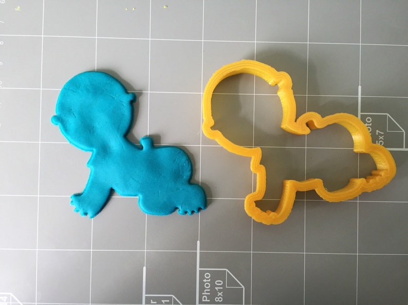 May include: A blue fondant baby cutout next to a yellow plastic cookie cutter in the shape of a baby. Both are on a gray grid surface.