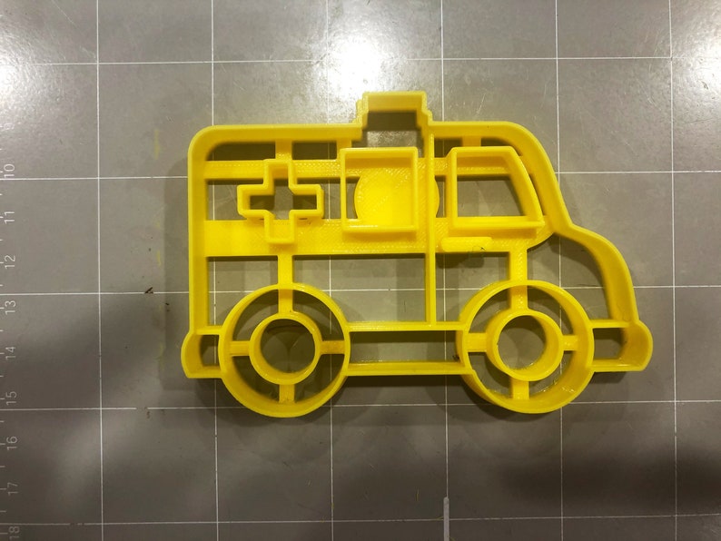 May include: Yellow plastic cookie cutter shaped like an ambulance. The cutter has a cross on the side and a window on the front.