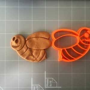 May include: A close-up of a brown clay bee cookie cutter and an orange plastic bee cookie cutter. The cookie cutters are on a gray grid surface.