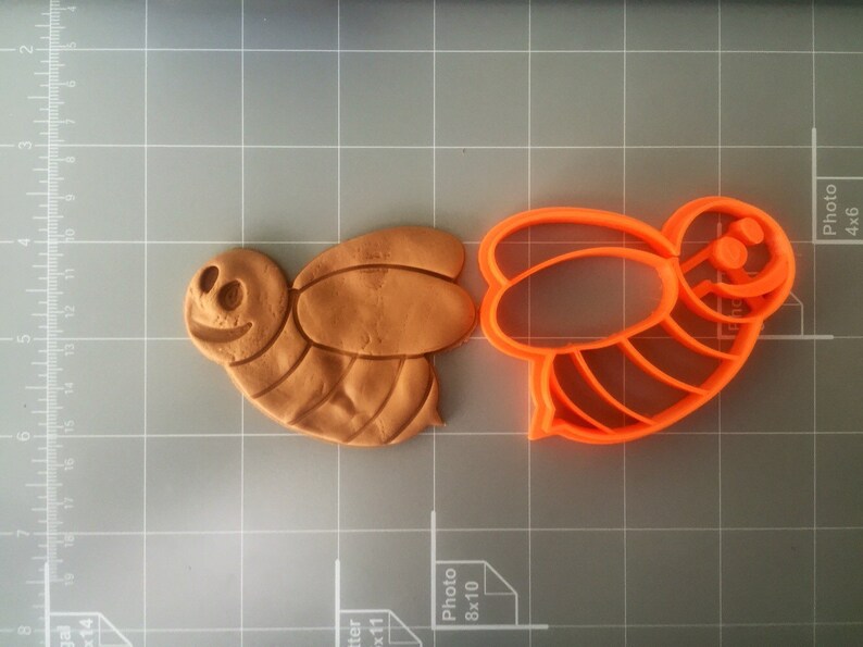 May include: A close-up of a brown clay bee cookie cutter and an orange plastic bee cookie cutter. The cookie cutters are on a gray grid surface.