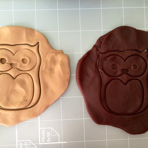 May include: Two owl-shaped cookie cutters, one brown and one dark brown, on a gray grid surface.