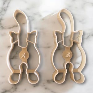 Può includere: Due formine per biscotti a forma di gatto beige. Ogni formina ha una coda, un corpo e delle zampe. Il design include un dettaglio "X" sul corpo. Le formine sono in plastica e sono progettate per la cottura.