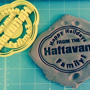 Puede incluir: Un cortador de galletas de plástico amarillo con el texto "Happy Holidays from the Haftavani Family!" y una galleta de arcilla marrón con el mismo texto impreso.