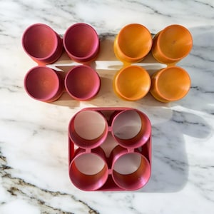 Puede incluir: Tres moldes de silicona para huevos, dos de color naranja rojizo y uno de color rojo oscuro. Los moldes tienen forma de círculo y tienen un borde elevado alrededor del borde. Los moldes están dispuestos en una fila sobre una superficie de mármol blanco.