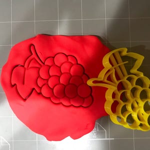 Puede incluir: Un cortador de galletas de plástico rojo y amarillo con forma de racimo de uvas. El cortador está sobre una superficie gris.