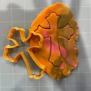 May include: A plastic cookie cutter in the shape of a cross with a decorative design. The cutter is orange and is placed on top of a piece of orange, pink, and brown play dough.