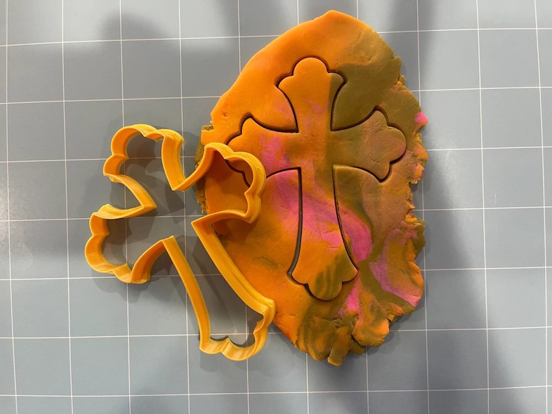 May include: A plastic cookie cutter in the shape of a cross with a decorative design. The cutter is orange and is placed on top of a piece of orange, pink, and brown play dough.