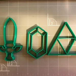 May include: Three green plastic cookie cutters shaped like a sword, a diamond, and a triangle. The triangle has a smaller triangle inside of it.