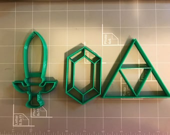 Puede incluir: Tres cortadores de galletas de plástico verde en forma de espada, diamante y triángulo. El triángulo tiene un triángulo más pequeño dentro.