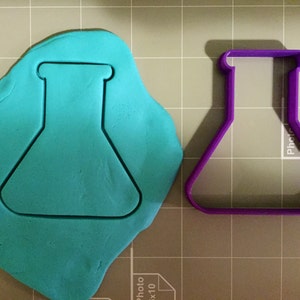 May include: A purple plastic cookie cutter shaped like a beaker. The cutter is on a gray and white grid surface. A blue piece of modeling clay has been pressed with the cutter.