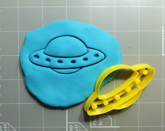 May include: A blue fondant imprint of a UFO with a yellow plastic cookie cutter of a UFO next to it.