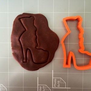 May include: A close-up of a red plastic cookie cutter shaped like a high-heeled shoe. The cookie cutter is on a grey grid surface. A brown piece of cookie dough is next to the cookie cutter.