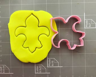 Könnte beinhalten: Ein gelber Fondant-Keksteig mit einem Ausstecher in Form einer Fleur-de-lis darauf. Rechts daneben ist ein rosa Plastik-Ausstecher in Form einer Fleur-de-lis zu sehen.