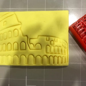 Puede incluir: Un molde de silicona amarillo con un diseño en relieve del Coliseo de Roma. El molde está al lado de un modelo del Coliseo impreso en 3D en rojo.