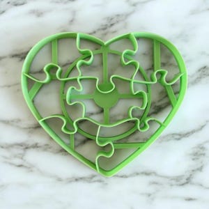 Puede incluir: Cortador de galletas de plástico verde en forma de corazón con un diseño de rompecabezas. El cortador tiene un círculo más pequeño en el centro rodeado de piezas de rompecabezas.